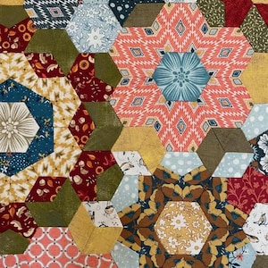 Può includere: Primo piano di una trapunta patchwork con un design floreale. La trapunta presenta una varietà di pezzi di tessuto esagonali nei toni del rosso, blu, verde e oro. Il motivo include motivi floreali e geometrici.