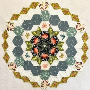 May include: A circular patchwork quilt with a floral design. The quilt features a central starburst pattern with red flowers and green leaves, surrounded by hexagonal patches in various colours and patterns, including white, green, and grey. The outer edge has a repeating floral border.