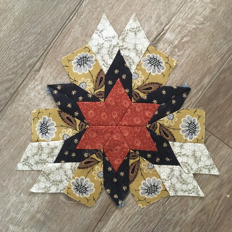 Peut inclure: Un bloc de courtepointe en forme d'&eacute;toile avec un sch&eacute;ma de couleurs rouge, noir et or. Le bloc est compos&eacute; de 12 pi&egrave;ces de tissu individuelles, chacune avec un motif diff&eacute;rent. Le centre du bloc est une &eacute;toile rouge avec un contour noir.
