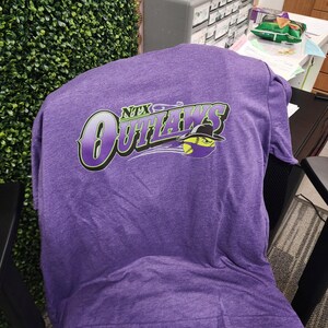 May include: A purple t-shirt with the text "NTX Outlaws" in a stylized font. The graphic includes a baseball wearing a hat. The shirt is made of a soft material and is likely for sports or team wear.