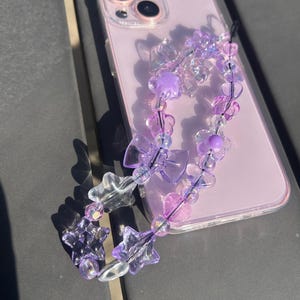 Puede incluir: Una funda de teléfono lavanda con un charm de teléfono con cuentas transparentes. El charm presenta cuentas transparentes y moradas en varias formas, incluyendo estrellas, flores y lazos. La funda del teléfono está sobre una superficie oscura.
