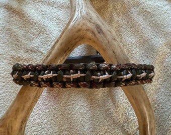 May include: A green, brown, and white paracord bracelet with a woven design. The bracelet is displayed on a white antler.