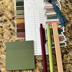 May include: A reading list printable with a stack of books bookmark, two pens, and a green sticky note. The printable has sections for title, date, and rating.