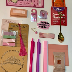 May include: A collection of reading-themed stationery and treats including a Dark Romance Book Club ticket, a honey packet, a Yogi tea bag, a gold spoon, two pens, a pink highlighter, sticky notes, a bookmark with a tassel, and a chocolate bar.