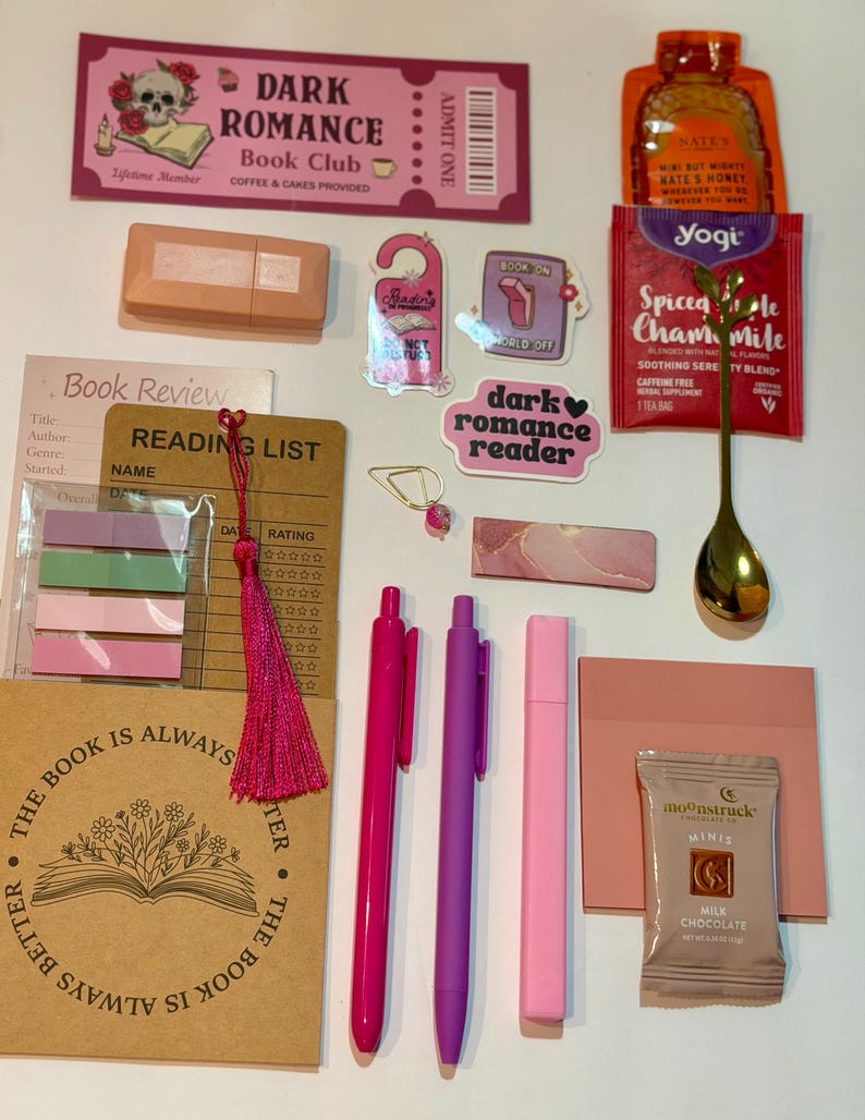 May include: A collection of reading-themed stationery and treats including a Dark Romance Book Club ticket, a honey packet, a Yogi tea bag, a gold spoon, two pens, a pink highlighter, sticky notes, a bookmark with a tassel, and a chocolate bar.