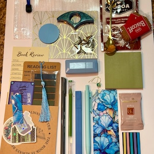 May include: A collection of stationery and treats including a clear zipper pouch, a Cocoa Classics cocoa mix packet, a Biscoff cookie, a gold spoon, a book review card, a reading list, pens, a floral bookmark, a milk chocolate bar, and stickers.