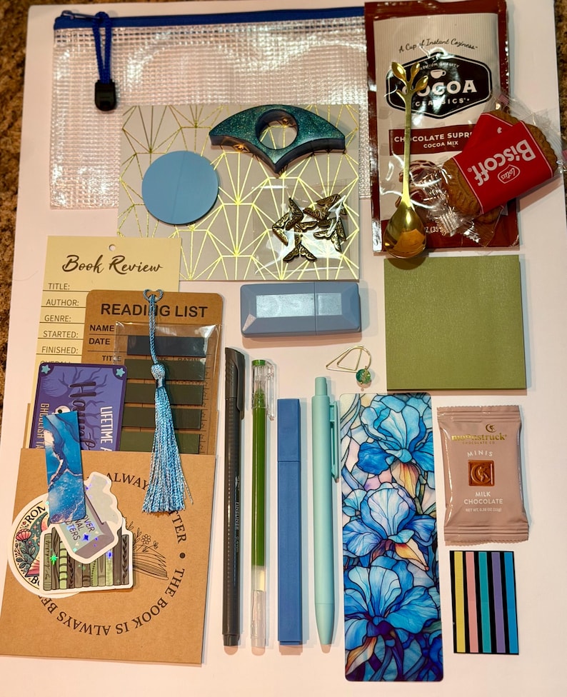 May include: A collection of stationery and treats including a clear zipper pouch, a Cocoa Classics cocoa mix packet, a Biscoff cookie, a gold spoon, a book review card, a reading list, pens, a floral bookmark, a milk chocolate bar, and stickers.