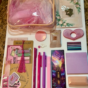 May include: A collection of stationery items including a clear pouch with pink tissue paper, a reading list card, a bookmark with a castle illustration, three pink pens, a pink highlighter, sticky notes, a chocolate bar, and a heart-shaped object.