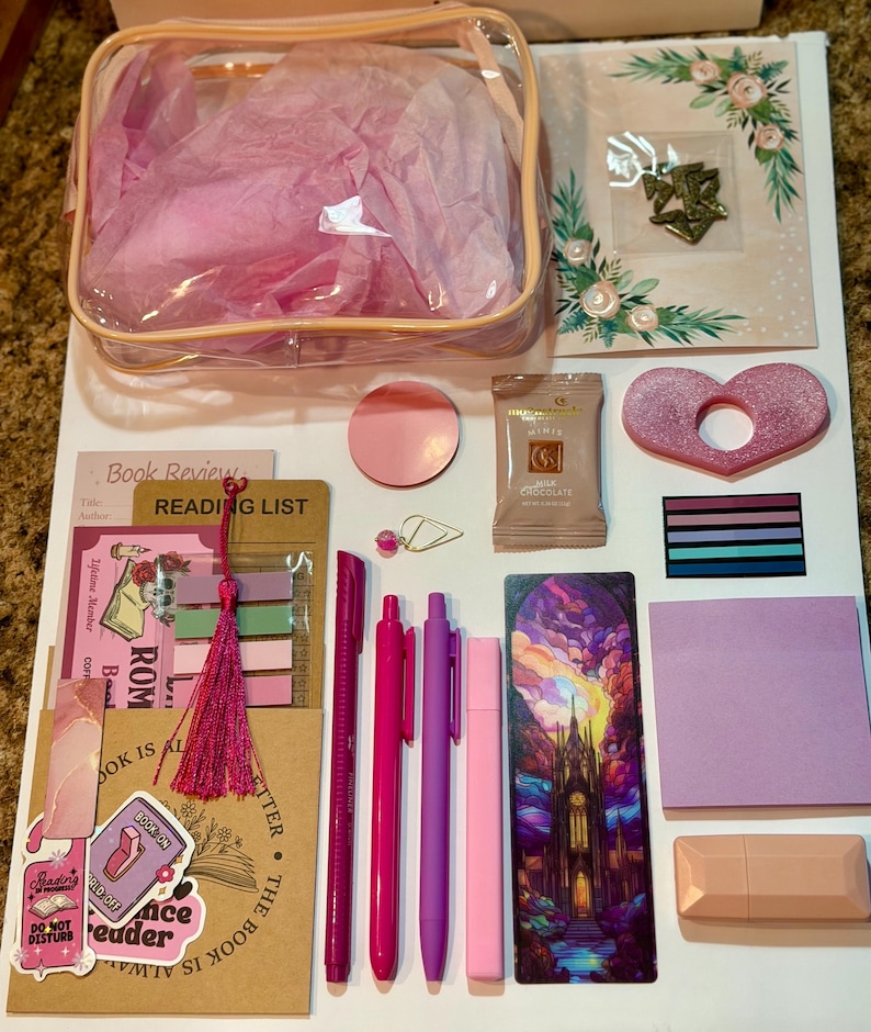 May include: A collection of stationery items including a clear pouch with pink tissue paper, a reading list card, a bookmark with a castle illustration, three pink pens, a pink highlighter, sticky notes, a chocolate bar, and a heart-shaped object.