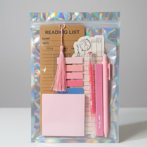May include: A stationery set in a holographic pouch containing a brown reading list notepad with a pink tassel, a stack of pink adhesive page markers, a square pink sticky note, a pink pen, a pink highlighter, and a white sticker with a globe.