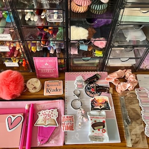 May include: A collection of stationery and accessories including a pink pencil case, a notepad, a reading log, stickers, a keychain, a pink scrunchie, a metal bookmark, a small candle, a tea bag, and a snack, all arranged on a wooden surface.