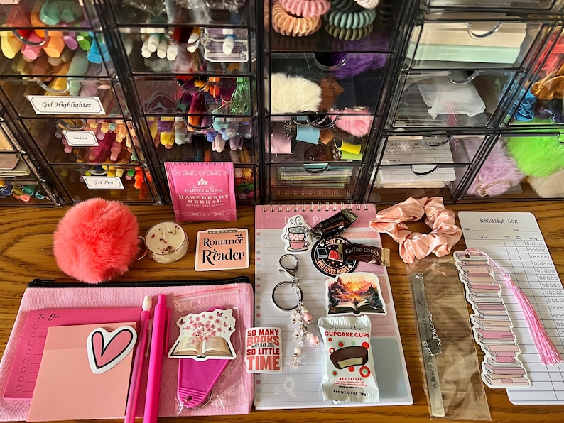 May include: A collection of stationery and accessories including a pink pencil case, a notepad, a reading log, stickers, a keychain, a pink scrunchie, a metal bookmark, a small candle, a tea bag, and a snack, all arranged on a wooden surface.