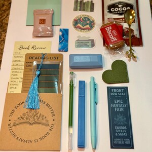 May include: A collection of stationery and reading accessories including a book review card, a reading list, a blue tassel, a green heart-shaped bookmark, a gold spoon, a Biscoff cookie, a cocoa packet, stickers, a blue highlighter, and pens.