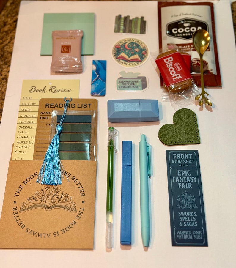 May include: A collection of stationery and reading accessories including a book review card, a reading list, a blue tassel, a green heart-shaped bookmark, a gold spoon, a Biscoff cookie, a cocoa packet, stickers, a blue highlighter, and pens.