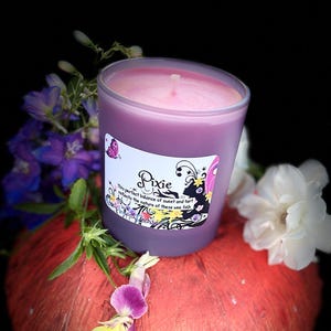 K&ouml;nnte beinhalten: Eine lila Kerze in einem transparenten Glasbeh&auml;lter mit dem Etikett "Pixie" steht auf einem roten, strukturierten Objekt. Die Kerze ist von lila und wei&szlig;en Blumen vor schwarzem Hintergrund umgeben.