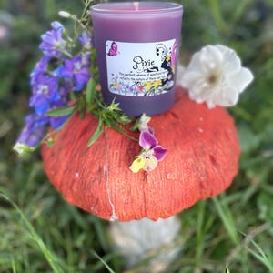 K&ouml;nnte beinhalten: Eine lila Kerze in einem Glas mit dem Etikett "Pixie" steht auf einem roten Pilz. Die Kerze ist von lila und wei&szlig;en Blumen umgeben, mit gr&uuml;nem Gras im Hintergrund. Das Etikett zeigt einen Schmetterling. Ein dekorativer Artikel.