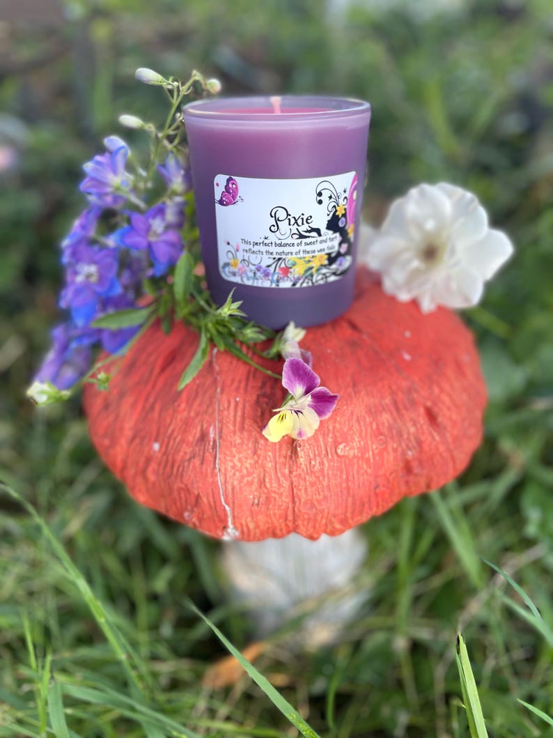 K&ouml;nnte beinhalten: Eine lila Kerze in einem Glas mit dem Etikett "Pixie" steht auf einem roten Pilz. Die Kerze ist von lila und wei&szlig;en Blumen umgeben, mit gr&uuml;nem Gras im Hintergrund. Das Etikett zeigt einen Schmetterling. Ein dekorativer Artikel.