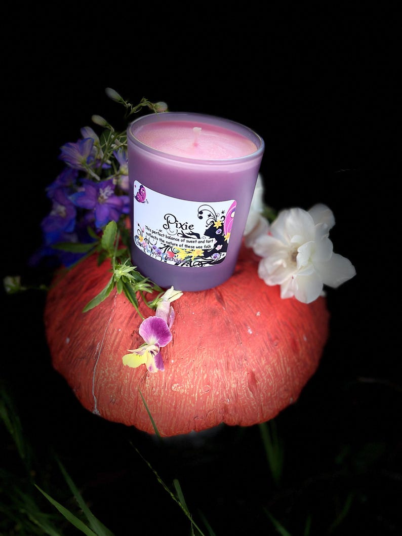 K&ouml;nnte beinhalten: Eine lila Kerze in einem transparenten Glasbeh&auml;lter mit dem Etikett "Pixie" steht auf einem roten, strukturierten Objekt. Die Kerze ist von lila und wei&szlig;en Blumen vor schwarzem Hintergrund umgeben.