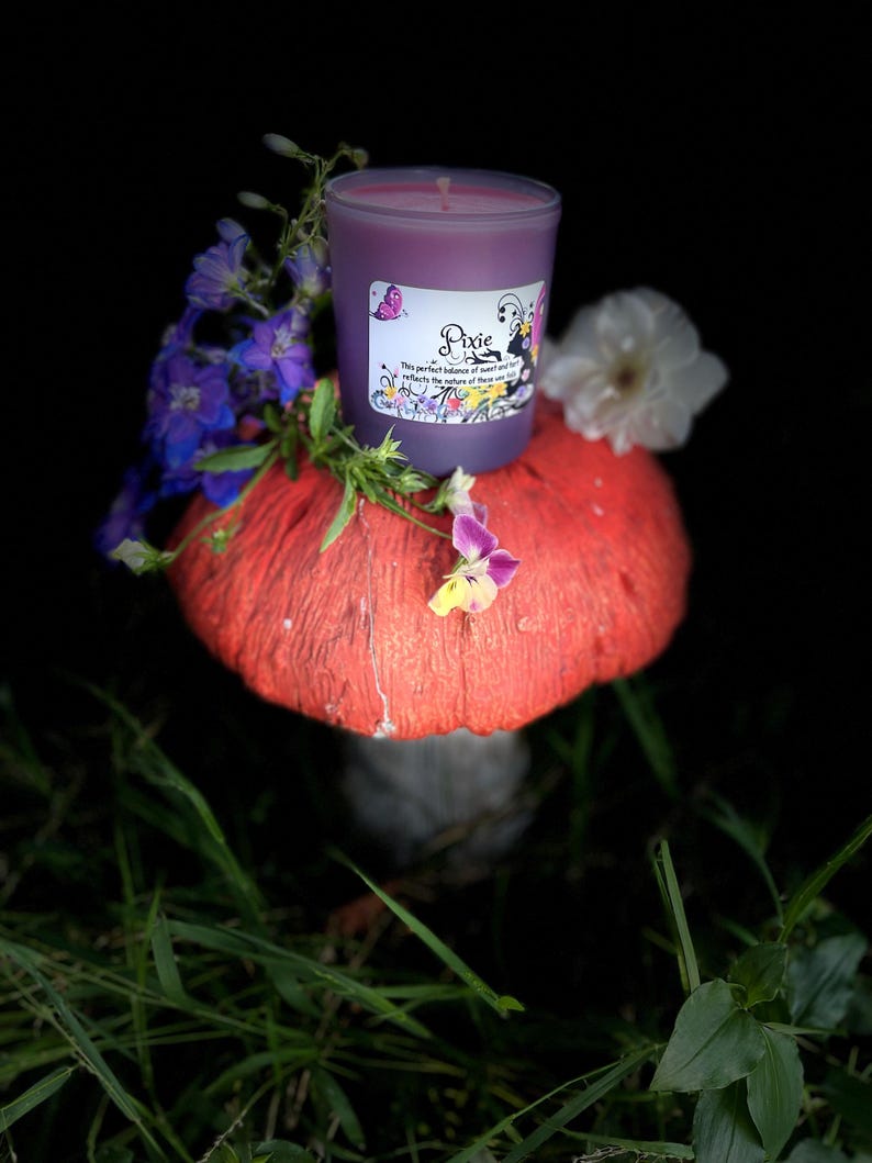 K&ouml;nnte beinhalten: Eine lila Kerze in einem Glasbeh&auml;lter mit dem Wort "Pixie" auf dem Etikett, steht auf einem roten Pilz. Die Kerze ist von lila und wei&szlig;en Blumen und gr&uuml;nem Gras umgeben. Das Kerzenetikett hat eine Schmetterlingsgrafik.
