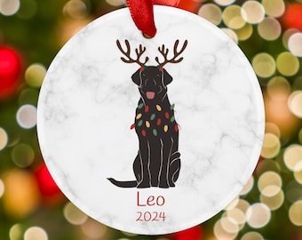 May include: A white marble Christmas ornament with a black Labrador Retriever wearing reindeer antlers and Christmas lights. The ornament has the name "Leo" and the year "2024" printed on it.
