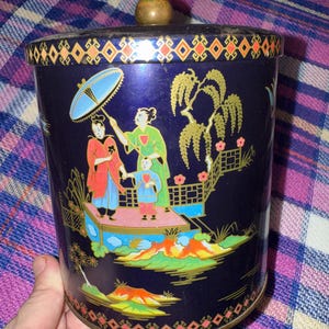May include: A vintage dark blue metal canister with a lid and a wooden knob. The canister features a colorful Asian-inspired design with figures, trees, and a bridge. The rim and base have a decorative geometric pattern.
