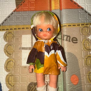 May include: A vintage doll with blonde hair, blue eyes, and freckles, wearing a colorful raincoat with a hood. The doll is dressed in a brown, yellow, and white patterned coat, white boots, and white socks. The doll is standing on a patterned surface.