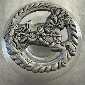May include: A clear glass jar with a silver-colored lid featuring a detailed rocking horse design. The lid has a circular shape with a braided edge, and the horse is in the center.