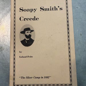 May include: A vintage book titled "Soapy Smith's Creede" by Leland Feitz. The cover is beige with black text and a portrait of a man in a hat. The bottom of the cover reads "The Silver Camp in 1892".