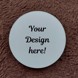 May include: A circular white coaster featuring the black text Your Design here! centered on the surface. The coaster has a smooth, flat finish and a clean, rounded edge, providing a blank space for custom graphics or personalized artwork.