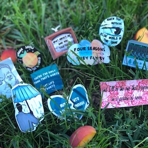 May include: Assortment of colorful stickers with various song lyrics and illustrations. The stickers feature a peach, a heart, a cloud, a television, an umbrella, and a heart with a broken line through it. The stickers are on a bed of green grass.
