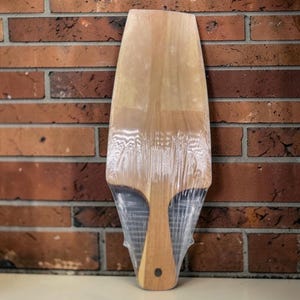 Könnte beinhalten: Ein helles Holzschneidebrett mit Griff, verpackt in klarem Kunststoff. Das Brett hat eine glatte Oberfläche und eine abgerundete Oberseite. Der Griff hat ein kleines, dunkel gefärbtes Loch. Der Hintergrund ist eine Backsteinmauer.