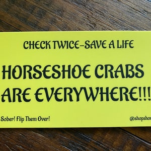 May include: Yellow sign with black text that reads "CHECK TWICE-SAVE A LIFE HORSESHOE CRABS ARE EVERYWHERE!!!!!" and "Drive Sober! Flip Them Over!" with the social media handle "@shopshoulderseason" at the bottom.