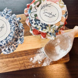 May include: Two glass jars with floral fabric covers. The jars are labeled "Sourdough Starter" and "Sourdough Discard". A wooden spoon with flour on it is in the foreground.