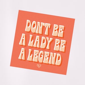 Może przedstawiać: Kwadratowa karta w kolorze brzoskwiniowym z tekstem "DON'T BE A LADY BE A LEGEND" w kolorze białym z efektem 3D. Tekst jest w retro stylu czcionki. W dolnej części karty znajduje się mały symbol nieskończoności.