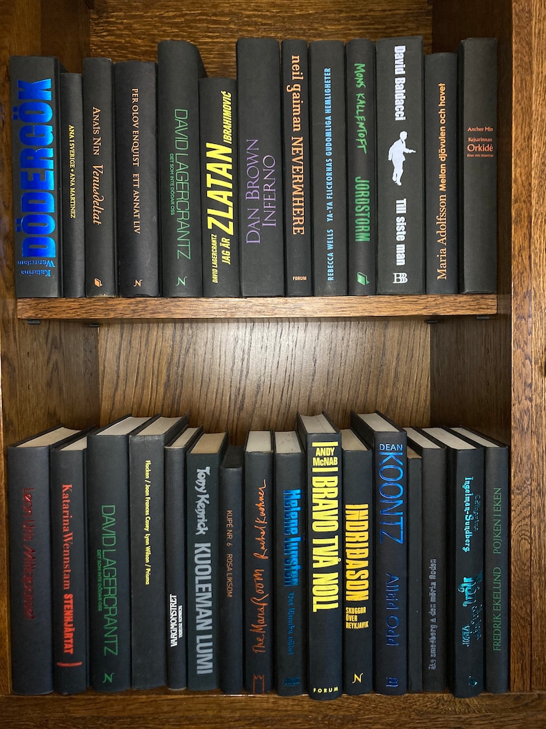 K&ouml;nnte beinhalten: Ein B&uuml;cherregal gef&uuml;llt mit schwarzen B&uuml;chern mit bunten Titeln. Die B&uuml;cher sind in zwei Reihen angeordnet. Die Titel beinhalten "Zlatan", "Inferno", "Neverwhere", "Jordsotm", "Till siste man", "Orkide", "KOONTZ", "Indri&eth;ason", "Bravo tv&aring; noll", "Kuoleman Lumi", "Stenhj&auml;rtat", "Vist", "Pojken i Eken" und mehr.