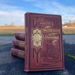 Peut inclure: Une pile de trois livres reliés anciens avec une couverture en cuir rouge et des lettres dorées. Le livre du haut est intitulé "Woodstock" par Walter Scott, Illustrated Uplaga, Stockholm, N.O. Gleerups Forlags Expeditions.