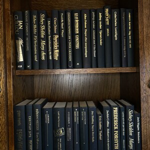 K&ouml;nnte beinhalten: Ein B&uuml;cherregal gef&uuml;llt mit schwarzen B&uuml;chern mit goldenen Lettern. Die B&uuml;cher sind nach Autor und Titel geordnet. Die B&uuml;cher sind alle in verschiedenen Gr&ouml;&szlig;en und Formen. Das B&uuml;cherregal ist aus Holz und hat eine dunkelbraune Oberfl&auml;che.