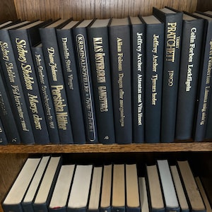 K&ouml;nnte beinhalten: Ein B&uuml;cherregal gef&uuml;llt mit schwarzen Hardcover-B&uuml;chern mit goldenen Lettern. Die Titel der B&uuml;cher beinhalten "Sidney Sheldon", "Klas &Ouml;stergren", "Allan Folsom", "Jeffrey Archer", "Terry Pratchett", "Colin Forbes" und "Danielle Steel".