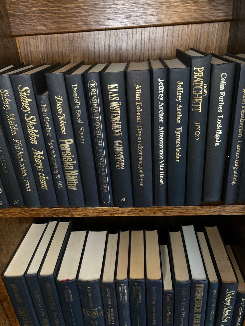 K&ouml;nnte beinhalten: Ein B&uuml;cherregal gef&uuml;llt mit schwarzen Hardcover-B&uuml;chern mit goldenen Lettern. Die Titel der B&uuml;cher beinhalten "Sidney Sheldon", "Klas &Ouml;stergren", "Allan Folsom", "Jeffrey Archer", "Terry Pratchett", "Colin Forbes" und "Danielle Steel".