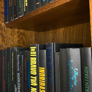 K&ouml;nnte beinhalten: Ein B&uuml;cherregal gef&uuml;llt mit schwarzen Hardcover-B&uuml;chern mit bunten Titeln. Die B&uuml;cher sind vertikal angeordnet und alle gleich gro&szlig;. Die Titel beinhalten "Bravo Two  Noli", "Indridason", "Koontz", "Catharina Ingelman-Sundberg" und "Pojken I Eken".