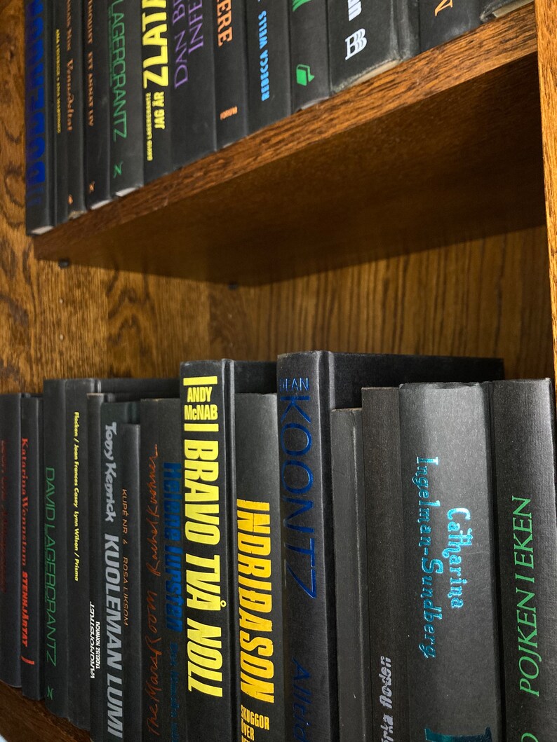K&ouml;nnte beinhalten: Ein B&uuml;cherregal gef&uuml;llt mit schwarzen Hardcover-B&uuml;chern mit bunten Titeln. Die B&uuml;cher sind vertikal angeordnet und alle gleich gro&szlig;. Die Titel beinhalten "Bravo Two  Noli", "Indridason", "Koontz", "Catharina Ingelman-Sundberg" und "Pojken I Eken".
