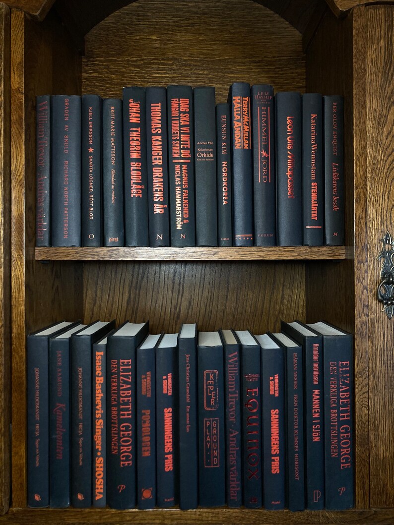 K&ouml;nnte beinhalten: Ein B&uuml;cherregal gef&uuml;llt mit schwarzen B&uuml;chern mit roten Titeln. Die B&uuml;cher sind in zwei Reihen angeordnet. Die obere Reihe hat Titel wie "Nordkorea", "Halla Andan", "Himmel Jord", "Stenhj&auml;rtat" und "Livl&auml;karens bes&ouml;k". Die untere Reihe hat Titel wie "Sanningens Pris", "Equinox", "Andras v&auml;rldar", "Mannen i sj&ouml;n" und "Den verkliga brottslingen".