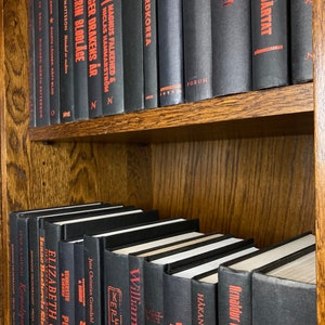 K&ouml;nnte beinhalten: Ein B&uuml;cherregal gef&uuml;llt mit schwarzen B&uuml;chern mit roten Titeln. Die B&uuml;cher sind in Reihen angeordnet und alle gleich gro&szlig;. Das B&uuml;cherregal ist aus Holz und hat eine dunkelbraune Oberfl&auml;che.