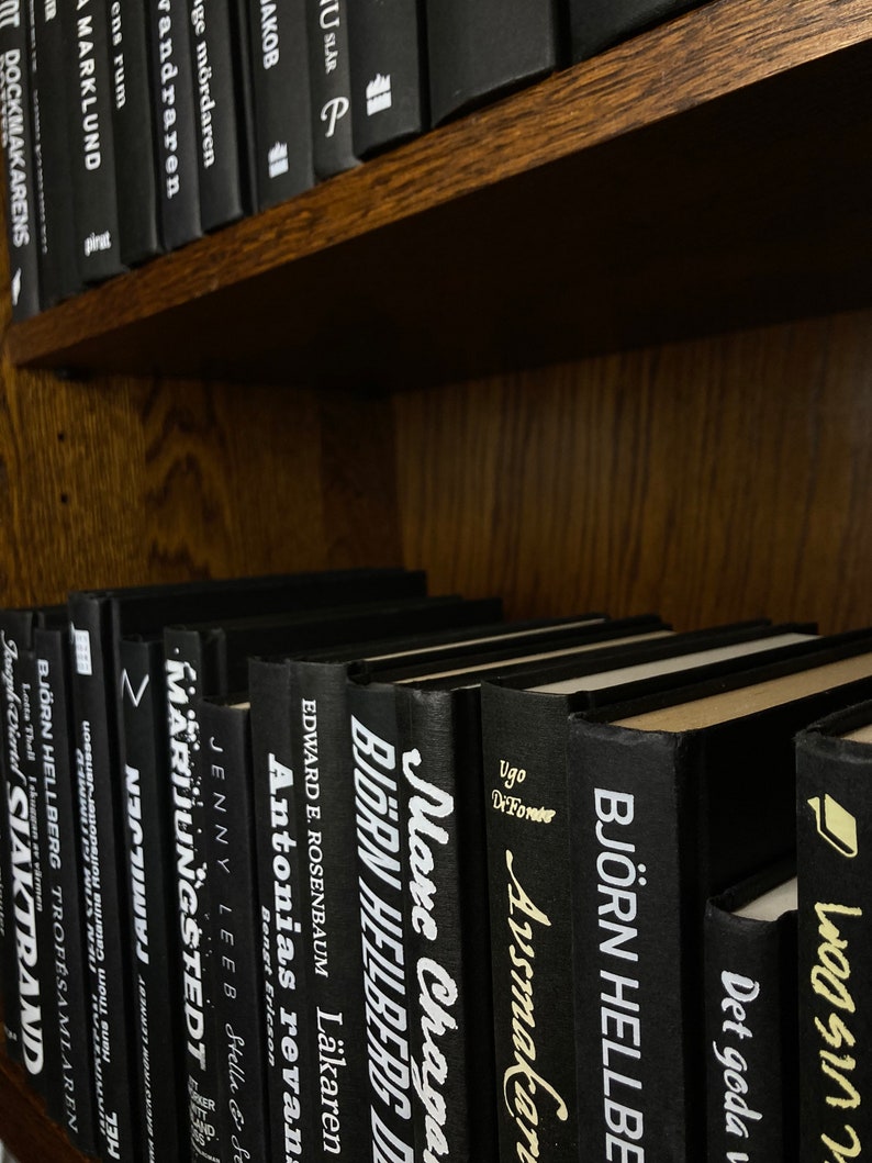 K&ouml;nnte beinhalten: Ein B&uuml;cherregal gef&uuml;llt mit schwarzen B&uuml;chern mit wei&szlig;en Texttiteln. Die Titel beinhalten "Antonias Revan", "Bjorn Heilberg", "Marc Chaga", "Avsmakaren", "Bjorn Hellberg", "Det goda", "Mardungstedt", "Sjaktrand" und mehr.