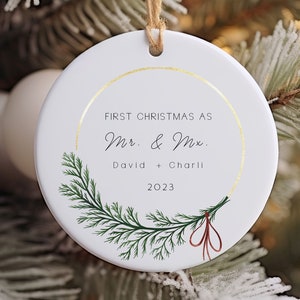 May include: White round Christmas bauble with a gold rim and a green pine branch design. The bauble says "First Christmas as Mr. & Mrs. David + Charli 2023".