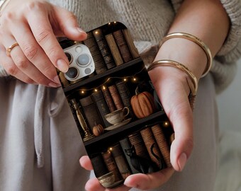 May include: A phone case featuring a bookshelf design with books, a pumpkin, and a teacup. The case has a dark background with warm lighting, creating a cozy, autumnal theme. The case is held by a person wearing a beige sweater and gold bracelets.