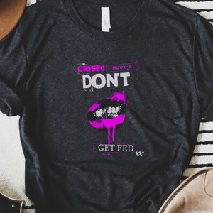 May include: A dark gray t-shirt with a pink graphic of a closed mouth with the text "Closed Mouths Don't Get Fed".