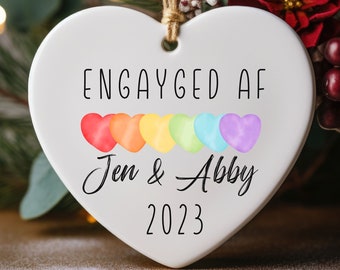 May include: White heart-shaped ceramic ornament with the text "Engayged AF" and a row of six rainbow-colored hearts. Below the hearts, the text reads "Jen & Abby 2023".
