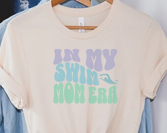 Könnte beinhalten: Ein hellbeiges T-Shirt mit einem grafischen Aufdruck, der "In My Swim Mom Era" in einer Retro-Schriftart mit einer Schwimmer-Silhouette zeigt. Der Text ist in den Farben Lila, Blau und Grün gehalten.