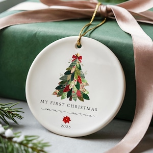 Może przedstawiać: Okrągła, biała ozdoba choinkowa z kolorowym wzorem choinki z patchworku, czerwoną kokardą oraz napisem "MY FIRST CHRISTMAS" i "emma marrie" z rokiem "2025". Ozdoba wisi na złotym sznurku, spoczywając na zapakowanym zielonym pudełku prezentowym z różową wstążką.